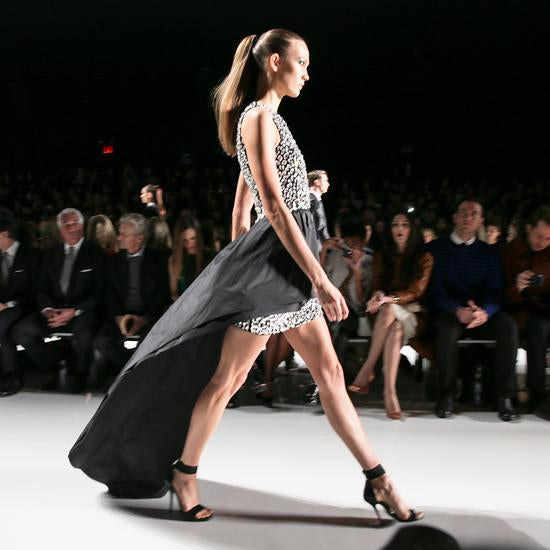 Model walking on a fashion show runway with an audience watching.