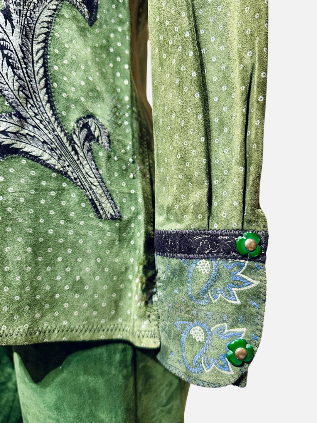 Close-up of a green embroidered garment, showing a sleeve with white dotted pattern, a blue and white floral cuff, and green decorative buttons. rare roberto cavalli for crissa suede 1970's appliqué painted collection flower snaps
