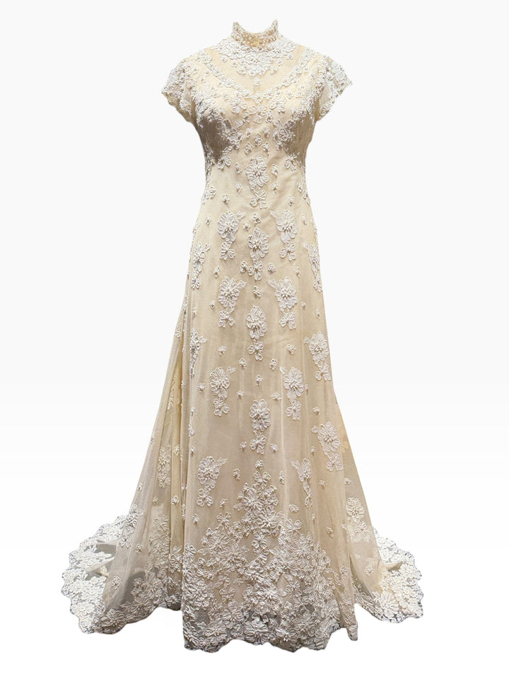 An elegant A-line wedding gown in champagne beige with intricate white floral lace appliques, a high mock neck, and short cap sleeves, shown against a plain white background.