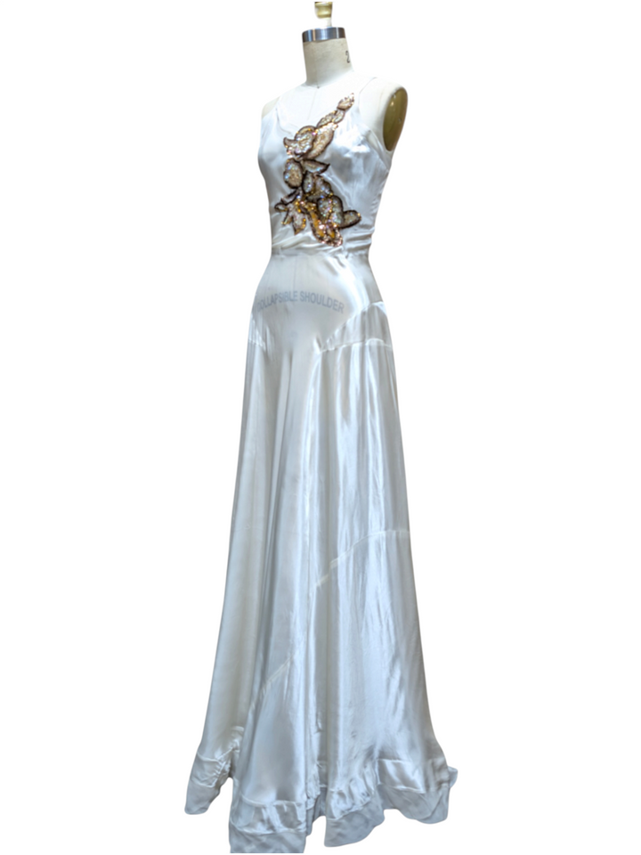 A sleeveless white satin maxi dress displayed on a mannequin against a plain white background. The dress features a fitted bodice with a gold and brown floral applique design on the chest and a ruffled hem.