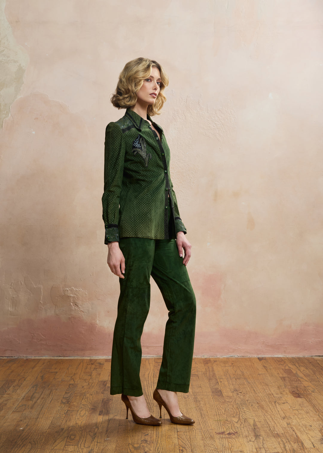 A woman models a dark green textured two-piece suit consisting of a long-sleeve button-up blazer with black embroidered details on the chest and matching wide-leg trousers, paired with brown high heels against a textured beige-pink wall and wooden floor.