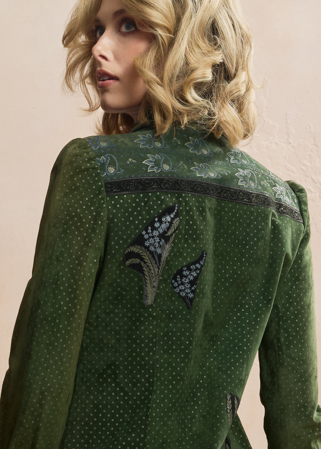 A close-up of a woman wearing a green embroidered jacket, showing the back of the garment with floral and paisley embroidery details against a neutral beige background. rare roberto cavalli for crissa suede 1970's appliqué painted collection