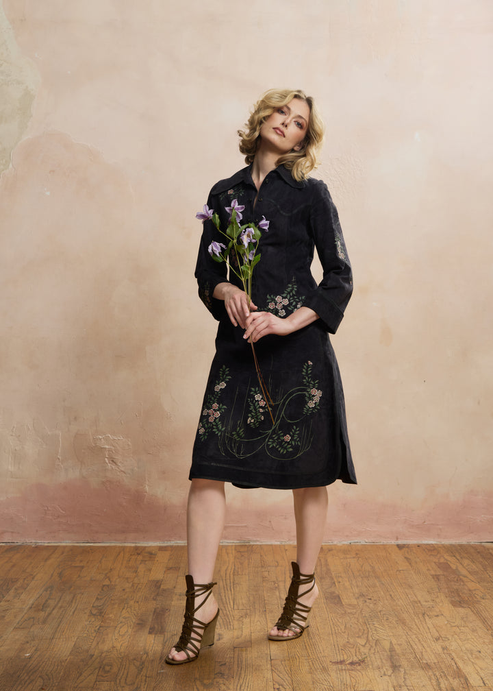 A woman models a black midi shirt dress with floral embroidery on the skirt, holding a small bouquet of purple flowers against a textured beige wall and wooden floor. ROBERTO CAVALLI RERACKED VOGUE 1970'S PAINTED SUEDE COLLECTION RARE - FASHION HISTORY MET MUSEUM