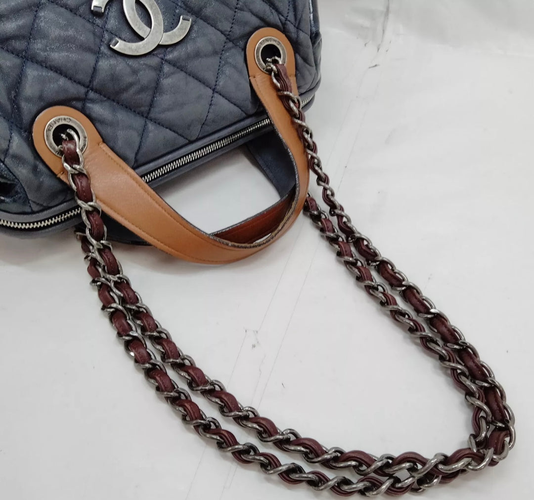 A close-up photo of a Chanel In The Mix tote bag in a vibrant blue color. The bag is made of quilted leather and features a chain-link strap and a gold Chanel logo on the front