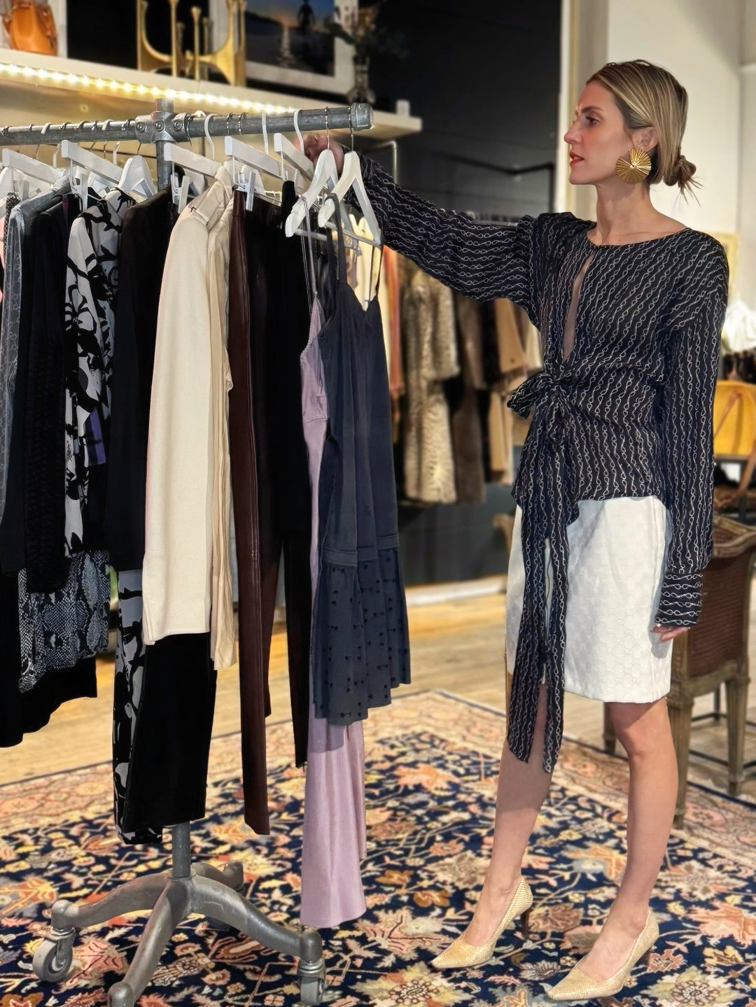 Woman standing in a clothing store with racks of clothes and a patterned rug on the floor.