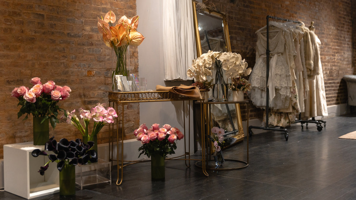 A boutique interior with a brick wall, dark wood floor, a gold-framed mirror, and a clothing rack holding cream-colored lace dresses. In the foreground, floral arrangements in pink, peach, and black are displayed on pedestals and a gold console table.
